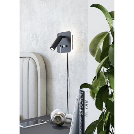 Fanzarotta 1 Square LED Wall Light in black, with a steel backplate and adjustable cylindrical reading spot, mounted above a bedside table beside a headboard and plant.