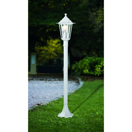 Fasvette Bollard Light in white aluminium, a traditional lantern-style post light with a six-sided glass head and finial on a slim pole, shown lit on a garden lawn at dusk.