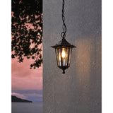 Fasvette lantern-style pendant light in black cast aluminium with clear glass panes, hanging from a black chain against an exterior wall at dusk.
