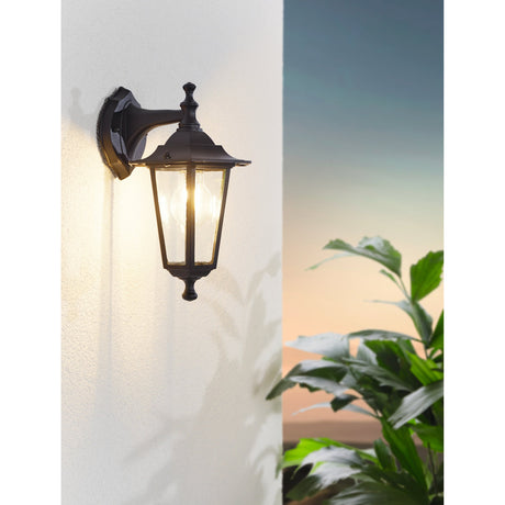 Fasvette downward outdoor wall light in black, a six-sided lantern with clear glass panels on a curved arm, mounted on an exterior wall beside greenery.