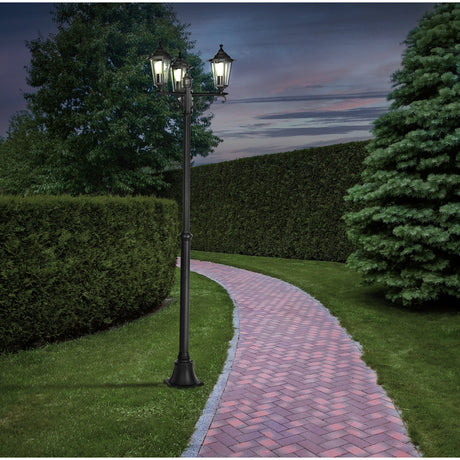 Fasvette three-light lamp post in black cast aluminium with hexagonal lanterns and scrolled arms, shown lit along a brick garden pathway at dusk.