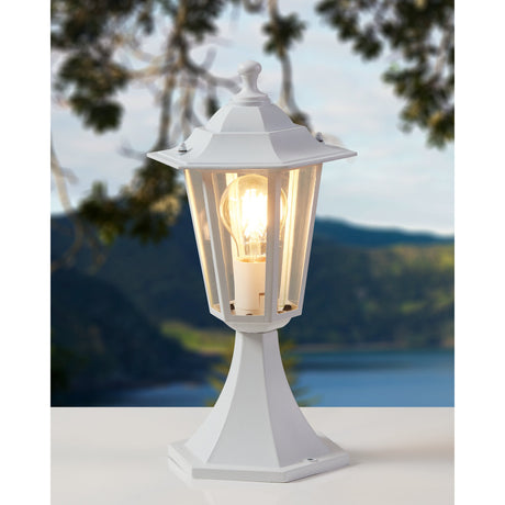 Fasvette outdoor post light in white, a hexagonal lantern with clear glass panels and finial on a tapered pedestal base, lit outdoors against a blurred lake and hillside backdrop.
