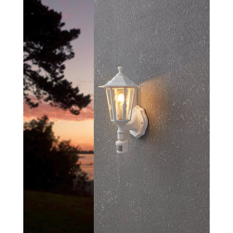 Fasvette white outdoor wall light with motion sensor, six-sided lantern with clear glass panels on a curved bracket, illuminated on an exterior wall at dusk.