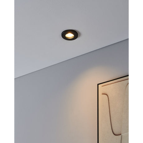 Favazzina 8.5cm round recessed downlight in black steel with clear glass cover, fitted in a sloped ceiling and casting warm light near a wall-mounted artwork.