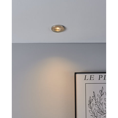 Favazzina 8.5cm recessed downlight in satin nickel with brushed steel bezel, shown fitted into a grey ceiling casting warm light across the wall beside a framed print.