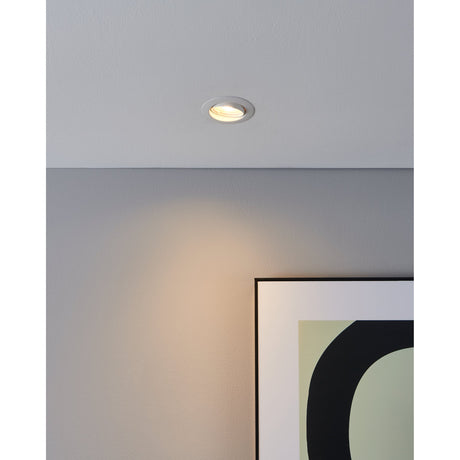 Favazzina 8.8cm round recessed downlight in white steel, shown installed in a ceiling casting a warm white glow onto a grey wall beside a framed print.