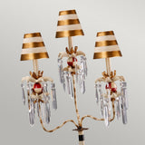 The Birdland 3 Arm Table Lamp - Cream & Gold features a classic candelabra-style design with three candle-like arms. Each arm is adorned with a striped gold and white lampshade, accented by red details and clusters of K9 cut glass, creating an elegant and timeless look.