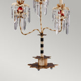 The Birdland 3 Arm Table Lamp in Cream & Gold showcases an elegant candelabra-style design with ornate detailing, featuring crystal-like pendants and red accents. It is adorned with K9 cut glass clusters, a striped central column, and rests on a star-shaped base against a plain light gray background.