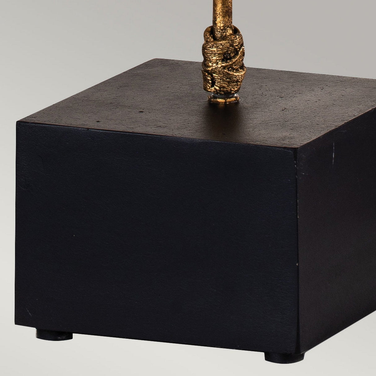 Close-up of the black cubic base supporting the Camilia 1 Light Table Lamp - Antique Gold & Black, featuring a gold metallic rod with a textured, wrapped design at the top. The plain light gray background accentuates its decorative element with subtle elegance.