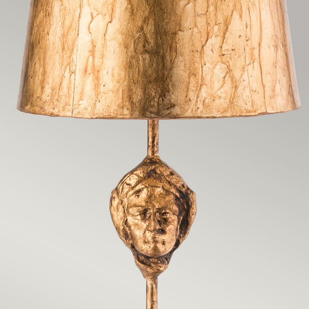 Introducing the Fragment 1 Light Table Lamp - Aged Gold, a piece that exudes vintage charm with its aged gold finish. It features a textured shade and a base decorated with an intricately sculpted face, crafting an elaborate artwork against a plain light gray background.