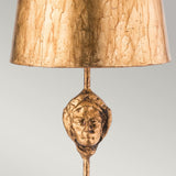 Introducing the Fragment 1 Light Table Lamp - Aged Gold, a piece that exudes vintage charm with its aged gold finish. It features a textured shade and a base decorated with an intricately sculpted face, crafting an elaborate artwork against a plain light gray background.