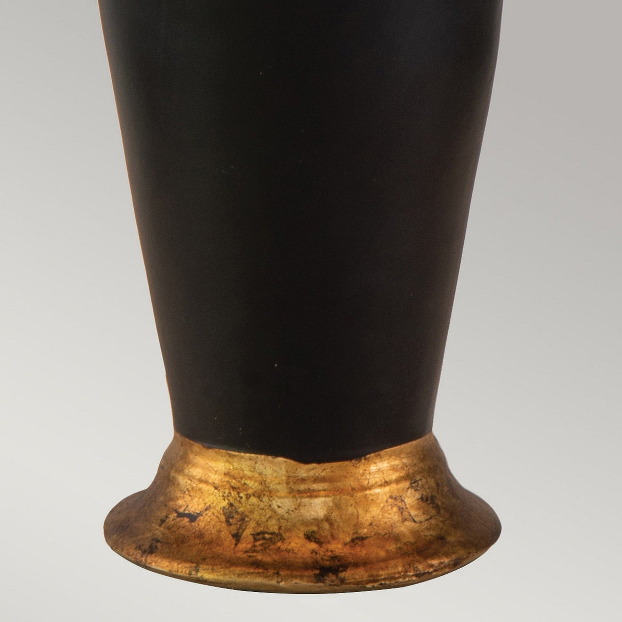A close-up of the Gallier 1 Light Table Lamp - Black & Gold reveals a slightly flared base with a textured, metallic black and gold finish. The background is plain and light-colored, accentuating the smooth and glossy surface of this striking piece of home decor.