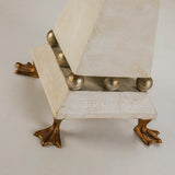 The Leda 1 Light Table Lamp, with its cream patina finish and gold leaf detailing, closely resembles a geometric sculpture. It features a stack of irregularly shaped white blocks accented by gold leaf spheres. The whimsical design is completed by two gold, duck-like feet extending from the base.