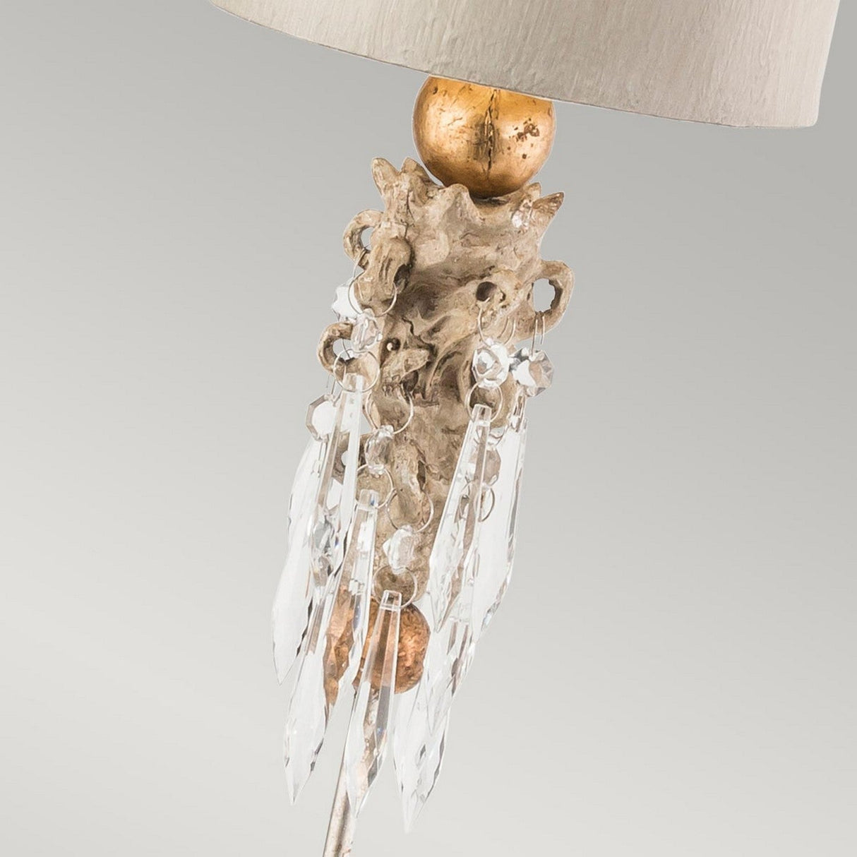The Madison 1 Light Table Lamp in Putty Patina and Gold Leaf presents an elegant design, featuring a decorative beige shade with an ornate, textured base adorned with K9 cut glass pendants, golden accents, and intricate metallic designs set against a plain gray background.