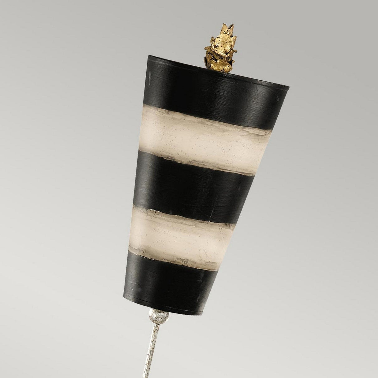 A Peony 1 Light Table Lamp in taupe and black stripes features a vertically oriented cone with alternating transparent and black bands, crowned with a small golden decorative piece. The stylish design is connected to a slender metallic rod and set against a plain gray background.