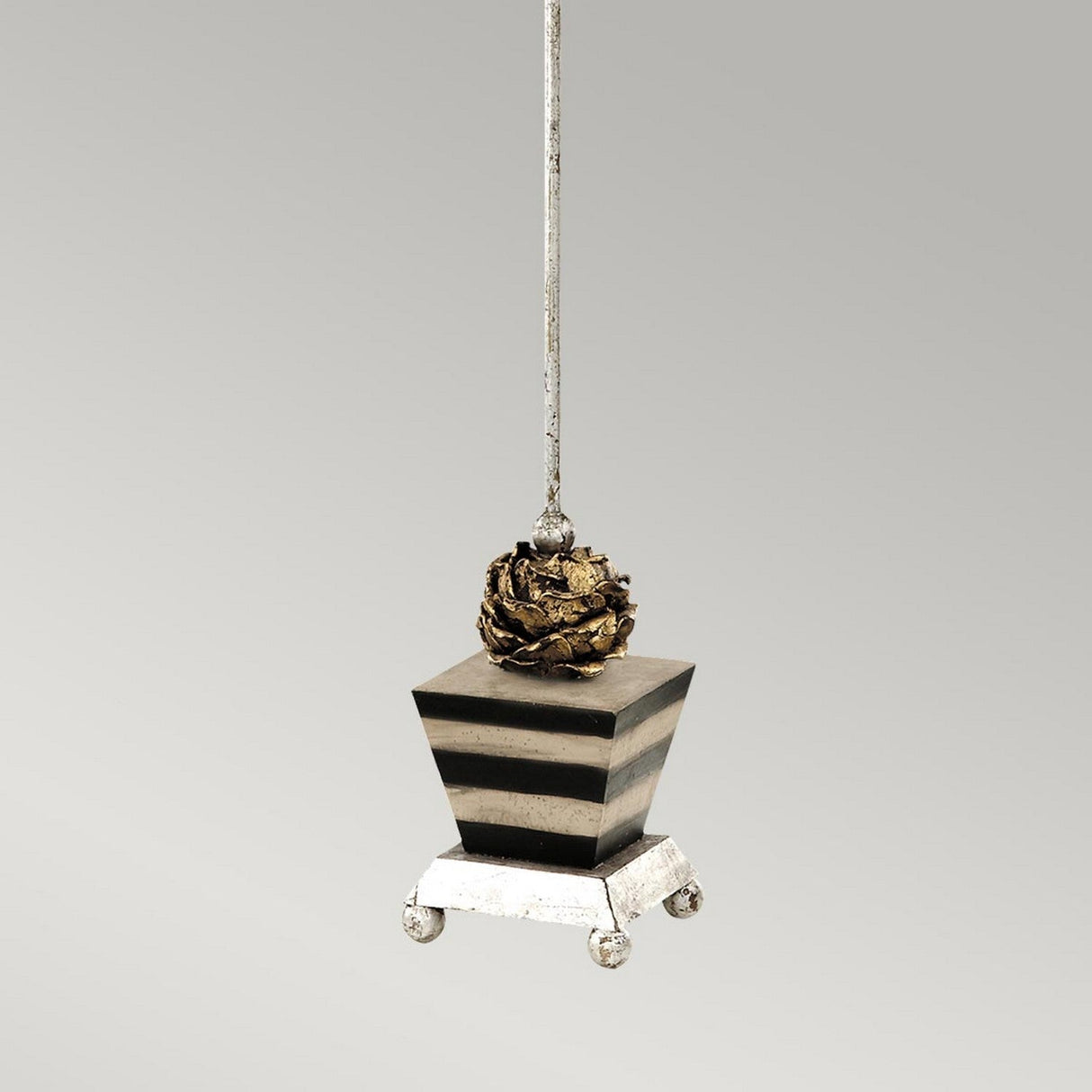 The Peony 1 Light Table Lamp, with its taupe and black striped pattern, showcases a square base suspended from a silver string. It features an intricate peony-like motif on a silver leaf finish and is supported by four small, spherical feet. The design contrasts beautifully with the plain gray gradient background.