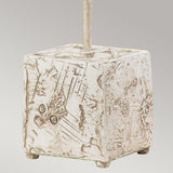 The Petra 1 Light Table Lamp - Bone White is characterized by its square stone base with abstract etching and an upward-extending metal rod. The surface boasts a weathered appearance adorned with geometric patterns, epitomizing contemporary design. Its plain, light gray backdrop accentuates the bone white base.
