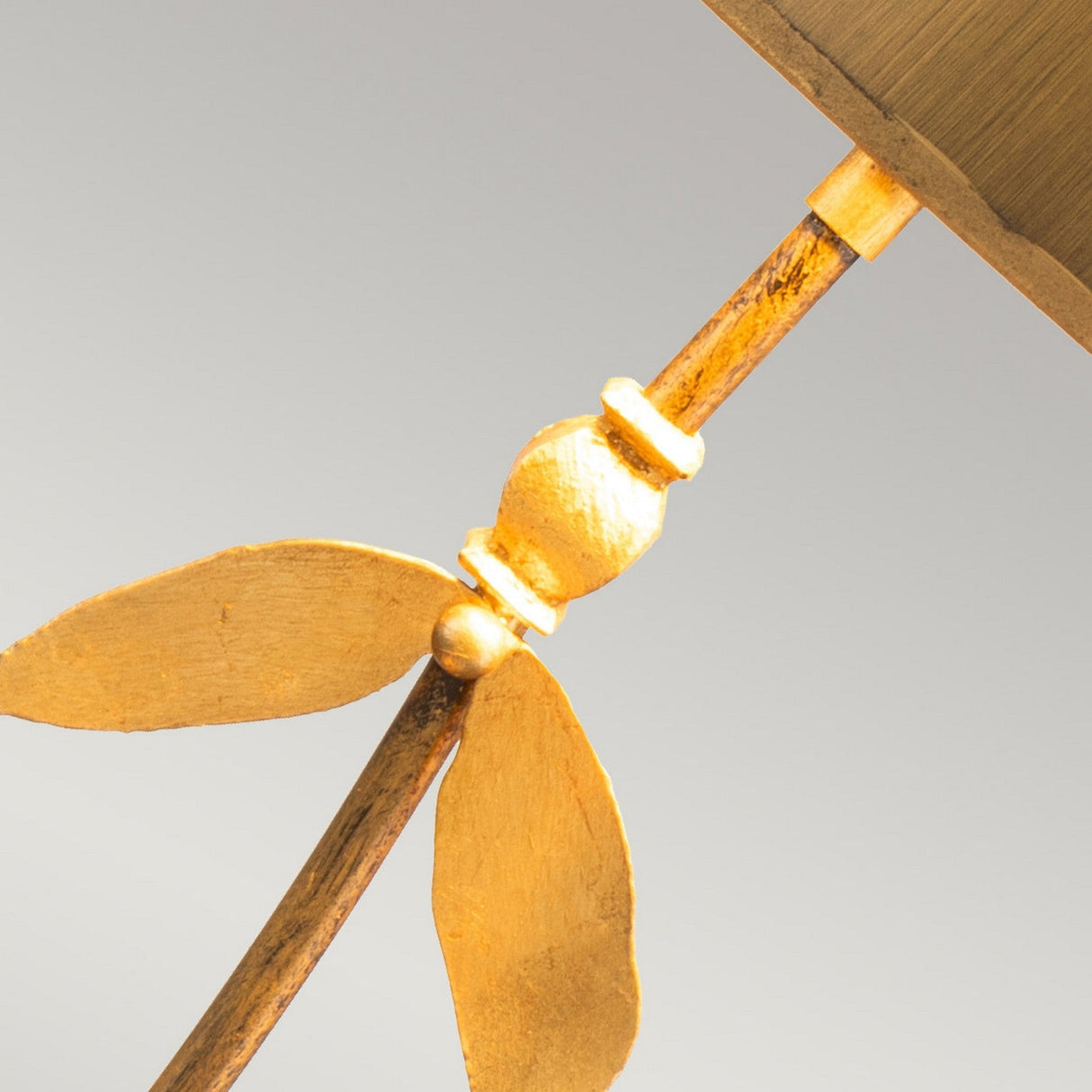A close-up of a decorative element from the Red Bell 1 Light Table Lamp - Gold Leaf features a stylized leaf design, highlighting nature-inspired sophistication with two symmetrical, elongated leaves attached to a vertical rod against a light gray background, showcasing the elegance of this Gold Leaf accent.