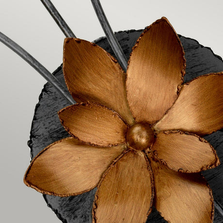 Close-up of the Simone 2 Light Wall Light, a decorative metallic gold flower sculpture with six petals set against a textured black oval background. This piece is crafted with gold leaf detailing and features two thin metallic rods partially visible, resembling an elegant wall fixture.