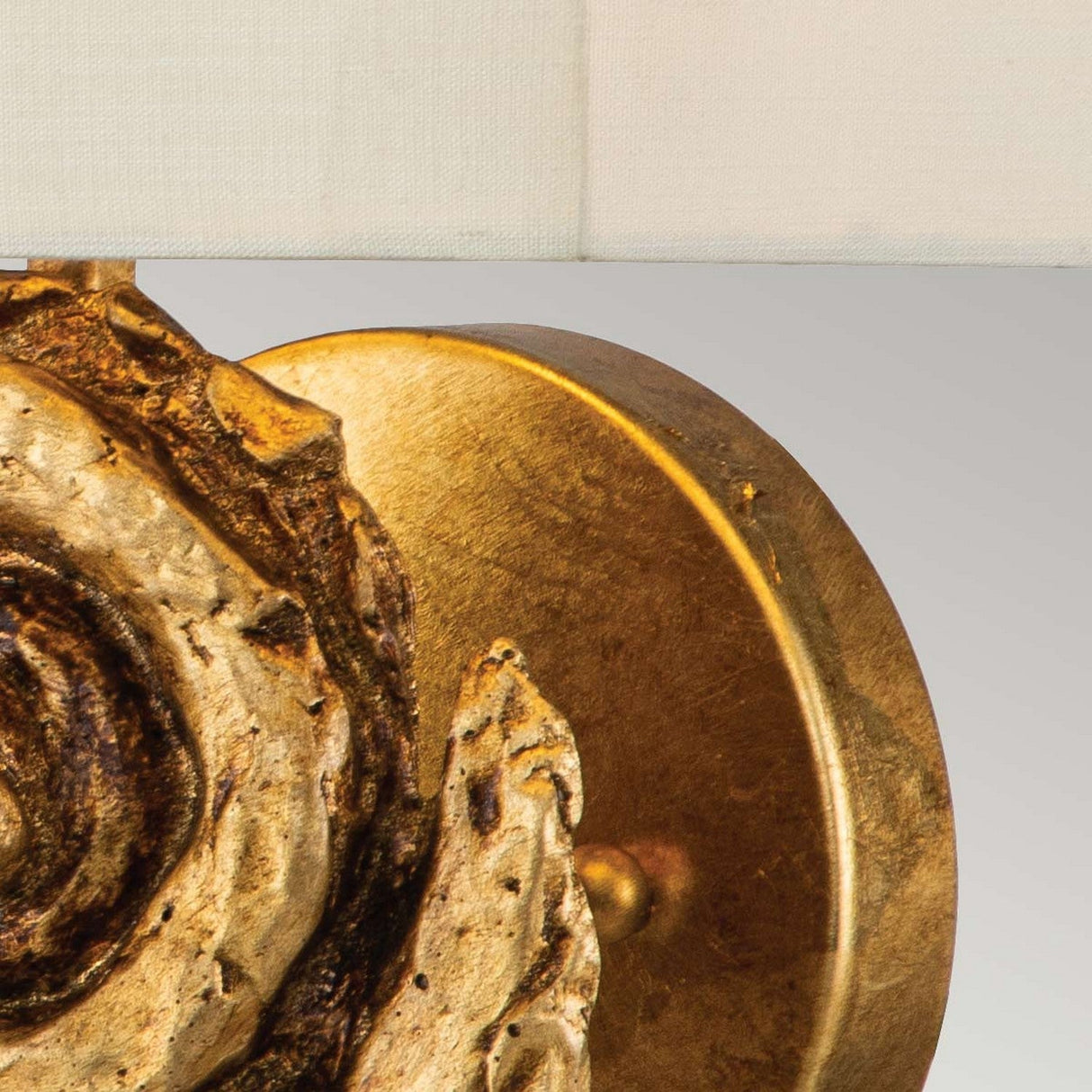 Close-up of a decorative Swirl 1 Light Large Wall Light - Gold Leaf, showcasing a textured spiral design on a round base. It is partially visible with a white fabric shade at the top, set against a light gray background that enhances the elegant gold leaf detailing.