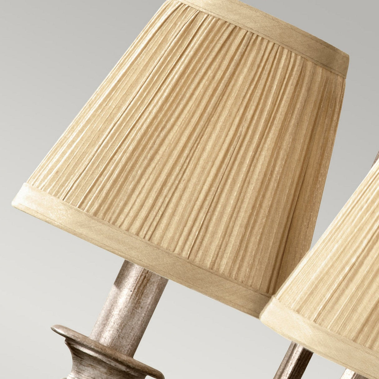 Close-up of the Abbey 3 Light Chandelier in a Silver Sand finish, featuring two beige pleated lamp shades with a textured design on metallic bases, set against a neutral background.