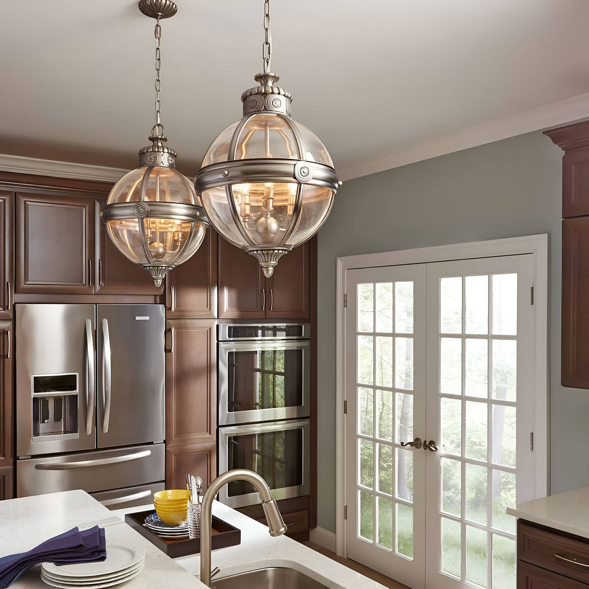 A contemporary kitchen showcases Victorian-inspired lighting with an Adams 3 Light Chandelier in Antique Nickel suspended above a marble island. Stainless steel appliances, such as a refrigerator and double wall oven, are seamlessly integrated into dark wooden cabinets. French doors offer a picturesque view of greenery.