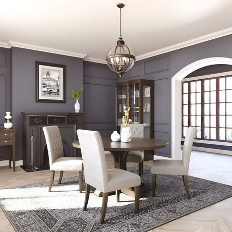 In a chic dining room, a round wooden table is accompanied by four beige upholstered chairs. A dark fireplace and cabinet provide contrast against the gray paneled wall. The space's Victorian-inspired elegance is highlighted by a large window and the Adams 4 Light Chandelier - Antique Nickel, with a patterned rug adorning the floor.