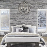 A cozy bedroom with a gray stone accent wall showcases the Allier 4 Light Chandelier in weathered oak wood and antique forged iron above. The bed is adorned with layers of white and gray bedding complemented by decorative pillows, while large windows offer a view of the snowy winter landscape outside.