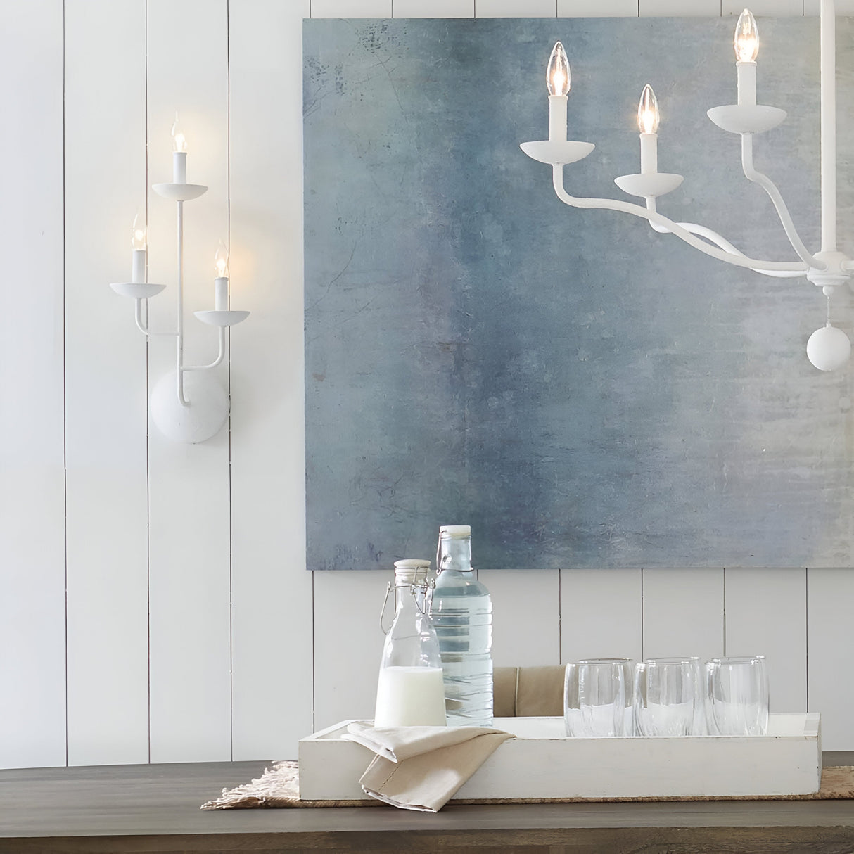 A modern dining area with walls in a textured plaster finish showcases the minimalist Annie 3 Light Wall Light in white. A large abstract blue painting complements the coastal-inspired design. Below, a table is adorned with a tray featuring glass bottles, glasses, and a milk bottle placed on a cloth napkin.