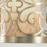 Close-up of the Arabesque 1 Light Wall Light, featuring an ornate metal design with swirling patterns and a flower-like motif, set against a beige fabric background. The metal showcases a silver leaf patina finish, offering a decorative and elegant appearance.