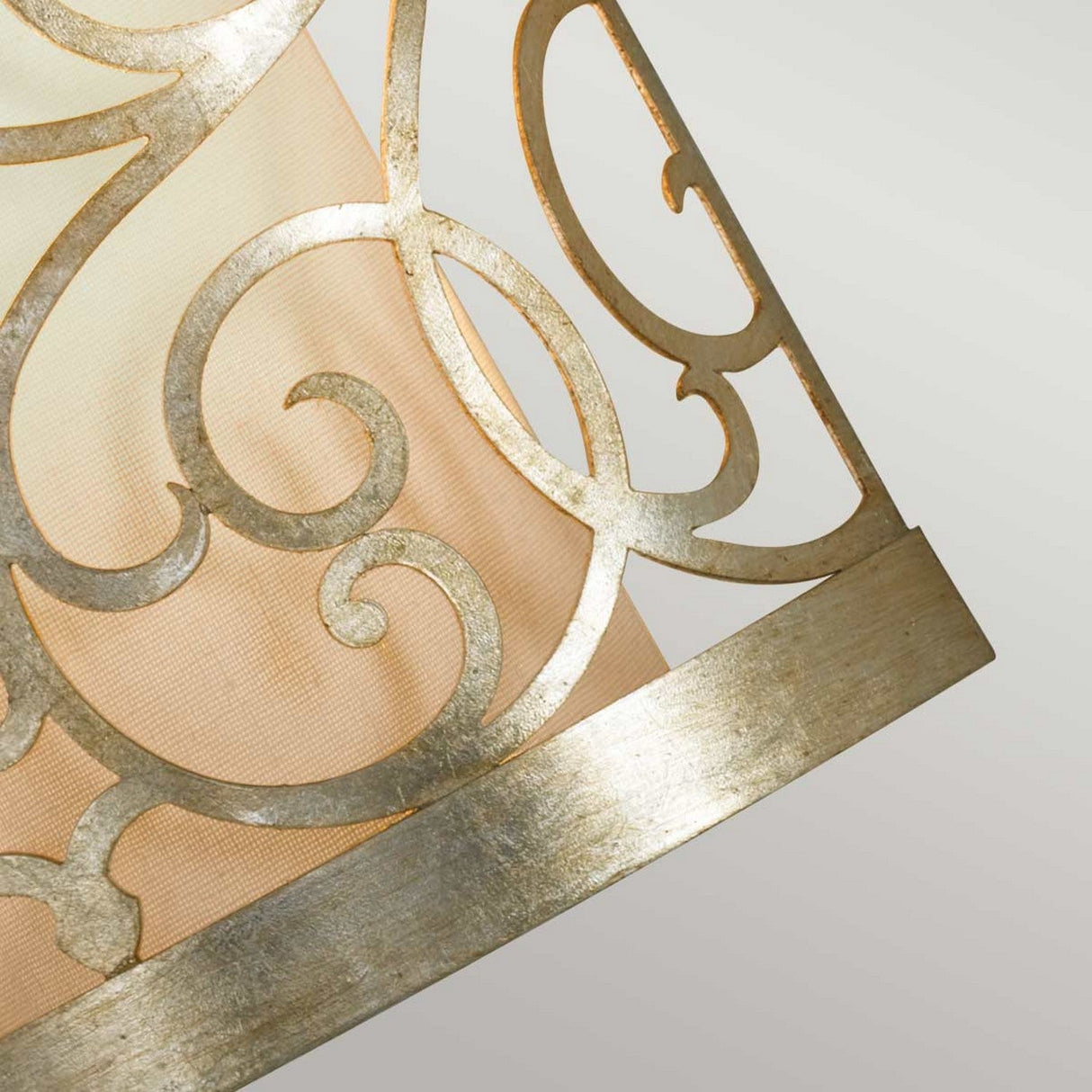 A close-up of the Arabesque 1 Light Wall Light in Silver Leaf Patina features a decorative metallic lampshade with laser-cut scrollwork showcasing intricate swirling patterns and a warm beige fabric lining, set against a light gray background.