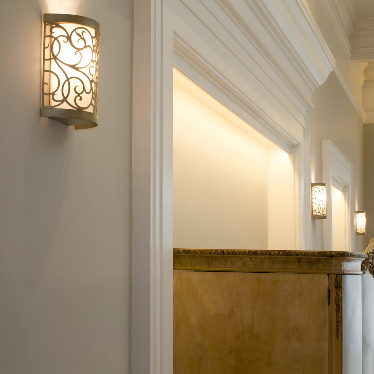 The Arabesque 1 Light Wall Light in Silver Leaf Patina casts a warm glow in a hallway featuring classic decor, complete with detailed moldings on the walls and ceiling. A wooden panel is partially visible, while the ornate light's laser-cut scrollwork enhances the space's cozy and elegant atmosphere.