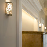 The Arabesque 1 Light Wall Light in Silver Leaf Patina casts a warm glow in a hallway featuring classic decor, complete with detailed moldings on the walls and ceiling. A wooden panel is partially visible, while the ornate light's laser-cut scrollwork enhances the space's cozy and elegant atmosphere.