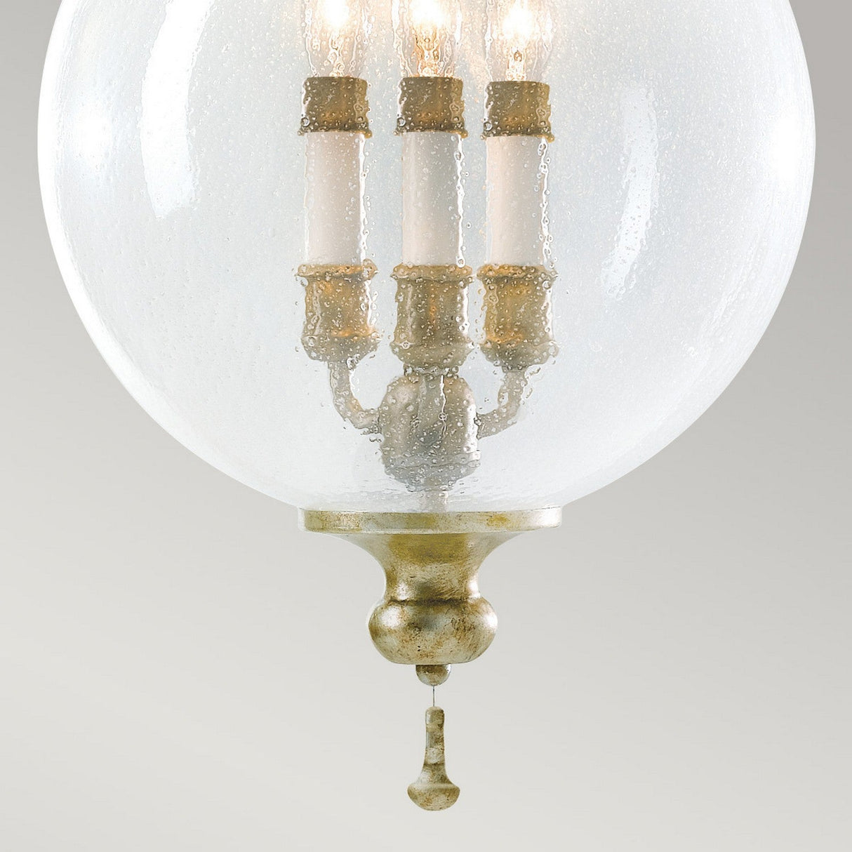 A close-up image of the Argento 3 Light Chandelier - Oxidized Silver Leaf showcases hand-blown bubble glass with a textured surface. Inside, three elegantly lit candles are housed in gold-tone holders, reminiscent of a mini chandelier. The fixture is finished with a decorative gold-tone finial at the base.