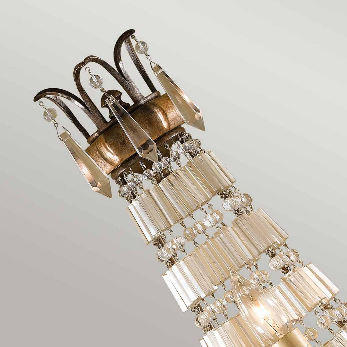 The Bellini 3 Light Wall Light in oxidized bronze/British bronze features a crown-shaped metal top and cascading rows of antique quartz crystals. Its Italian-inspired design reflects light beautifully, resulting in a shimmering effect against a light gray background.