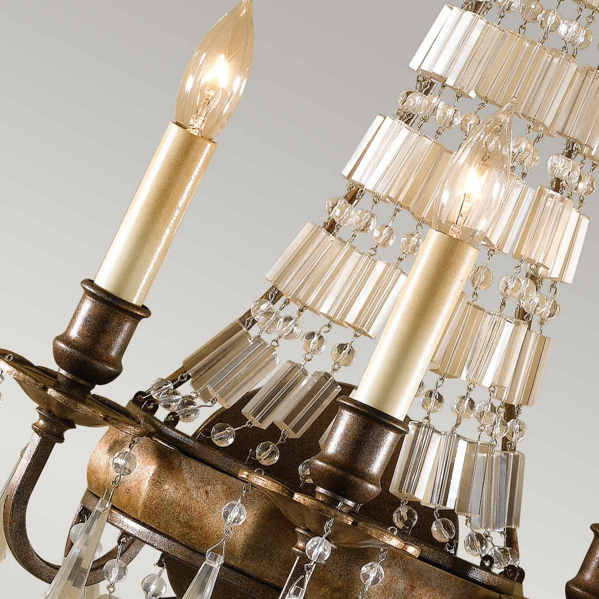 Close-up of the Bellini 3 Light Wall Light, featuring an oxidized bronze finish with three lit candle-shaped bulbs. The wall light showcases intricate metalwork and cascading crystal prisms, creating a sparkling effect against a soft gray background.