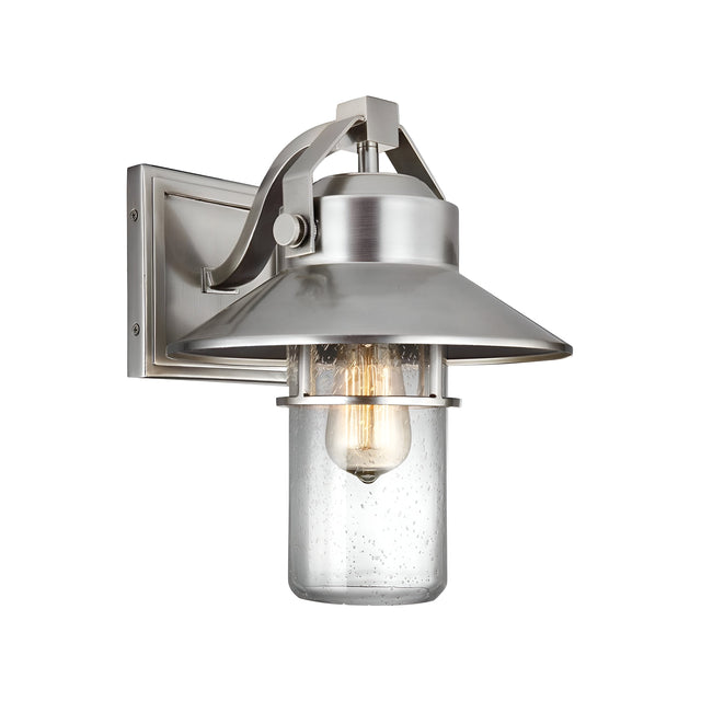 The Boynton 1 Light Outdoor Medium Wall Lantern - Painted Brushed Steel features a modern wall-mounted sconce design with a brushed metal finish and a clear glass cover. Its visible light bulb adds an industrial touch, while the weather-resistant lantern includes a rectangular backplate and is slightly angled downward.