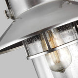 Close-up of the Boynton 1 Light Outdoor Medium Wall Lantern in Painted Brushed Steel, showcasing a metallic, weather-resistant design with a clear, textured glass enclosure. Inside, an Edison-style bulb emits a warm glow. This sleek fixture adds modern charm against its plain gray background.