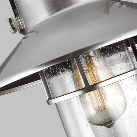 Close-up of the Boynton 1 Light Outdoor Medium Wall Lantern in Painted Brushed Steel, showcasing a metallic, weather-resistant design with a clear, textured glass enclosure. Inside, an Edison-style bulb emits a warm glow. This sleek fixture adds modern charm against its plain gray background.