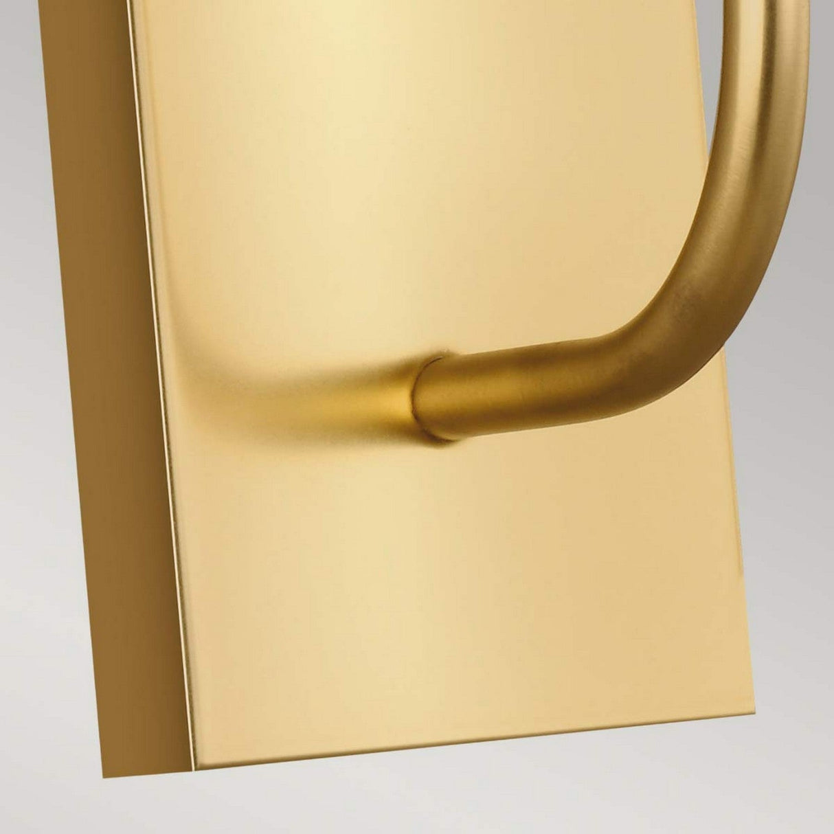 A close-up showcases a burnished brass door handle affixed to a rectangular gold plate, set against a light gray backdrop. This design embodies a sleek, contemporary aesthetic with smooth curves and a polished finish, echoing the elegance of the Brianna 1 Light Wall Light in Burnished Brass.