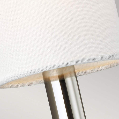 A close-up of the Brianna 1 Light Wall Light reveals its white fabric shade paired with a sleek, polished nickel base. The lamp radiates contemporary elegance, emphasizing the seamless connection between its shade and base, while ambient lighting enhances the neutral, light gray backdrop.