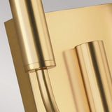 A close-up of the Brianna 2 Light Wall Light in burnished brass showcases its modern design with a dual-light configuration, featuring cylindrical and rectangular elements against a light background. The sleek, reflective surfaces, enhanced by White Linen cylindrical shades, add to its elegant allure.