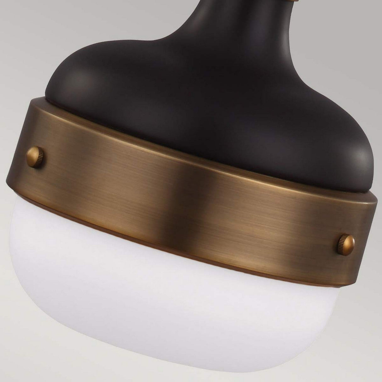 A close-up of the Cadence 1 Light Mini Pendant showcases its black and bronze body with a white frosted glass shade. This fixture embodies an industrial lighting style, featuring decorative screws that secure a bronze band around the glass for a vintage-industrial feel.