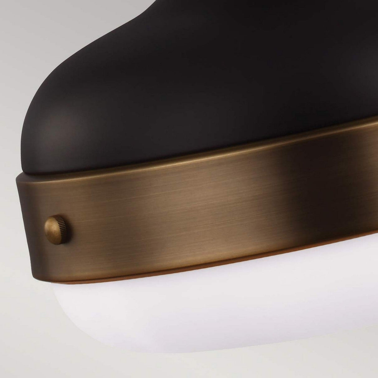 Close-up of the Cadence 2 Light Pendant showcasing a smooth, matte black top paired with a dark antique brass band and accented by a small screw. The bottom features a white, frosted glass diffuser, merging industrial lighting elements into a sleek, modern design set against a neutral background.