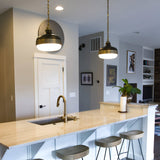 The modern kitchen features a marble island countertop accompanied by four wooden stools. Industrial pendant lighting, specifically the Cadence 2 Light Pendant in Dark Antique Brass/Matte Black, elegantly hangs from the ceiling, adding a touch of charm. A single-handle faucet is installed on the island. Wall art and a plant decorate the space, which opens up to a living area in the background.