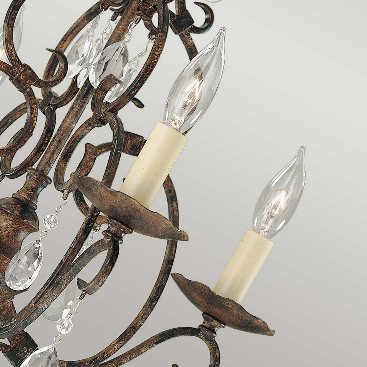 The Chateau 4 Light Chandelier in Mocha Bronze showcases two candle-style incandescent bulbs, ornate filigree, and hanging crystal accents against a grey backdrop.