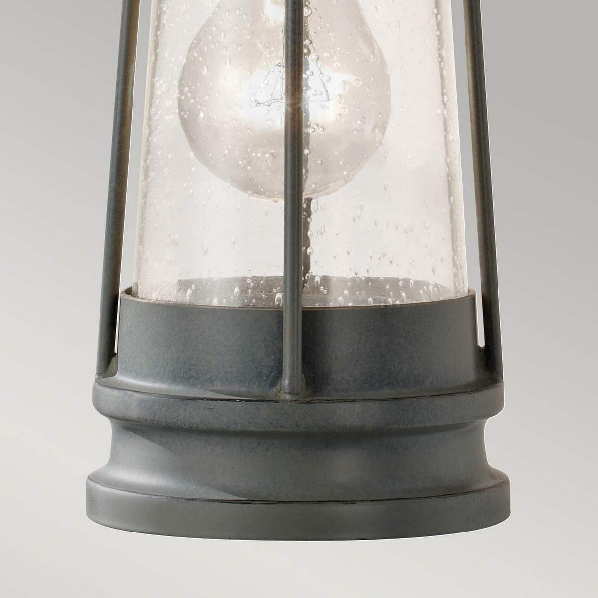 Close-up of the Chelsea Harbor 1 Light Outdoor Wall Lantern in Storm Cloud, featuring a vintage aesthetic with a clear glass cover and visible light bulb inside. The lantern showcases a weathered finish, cylindrical shape with vertical metal supports, and a round base, set against a neutral gray background.