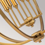 The Corinne 6 Light Chandelier in Burnished Brass is highlighted in a close-up, revealing its mechanical structure with gears and parallel rods. The intricate design of this chandelier showcases metallic finishes and cylindrical elements arranged in a curved framework, evoking the silhouette of an open orb against a light background.