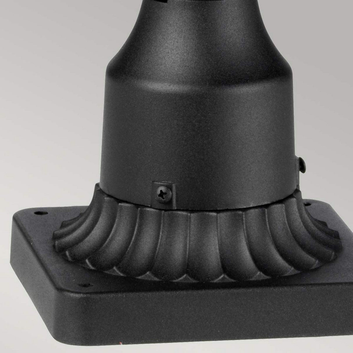Close-up of the Cotswold Lane 3 Light Outdoor Large Pedestal in black, showcasing its decorative fluted design. The square metal base includes a small screw near the curved section, ideal for an outdoor pedestal light. The light gray background enhances the stunning black finish of this lighting piece beautifully.