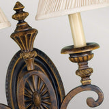 Close-up of the Drawing-1 Room 2 Light Wall Light in a vintage style. The fixture, with a walnut finish reminiscent of Edwardian design, features two candle-like lights topped with pleated beige shades. Intricate carvings enhance its circular medallion base.